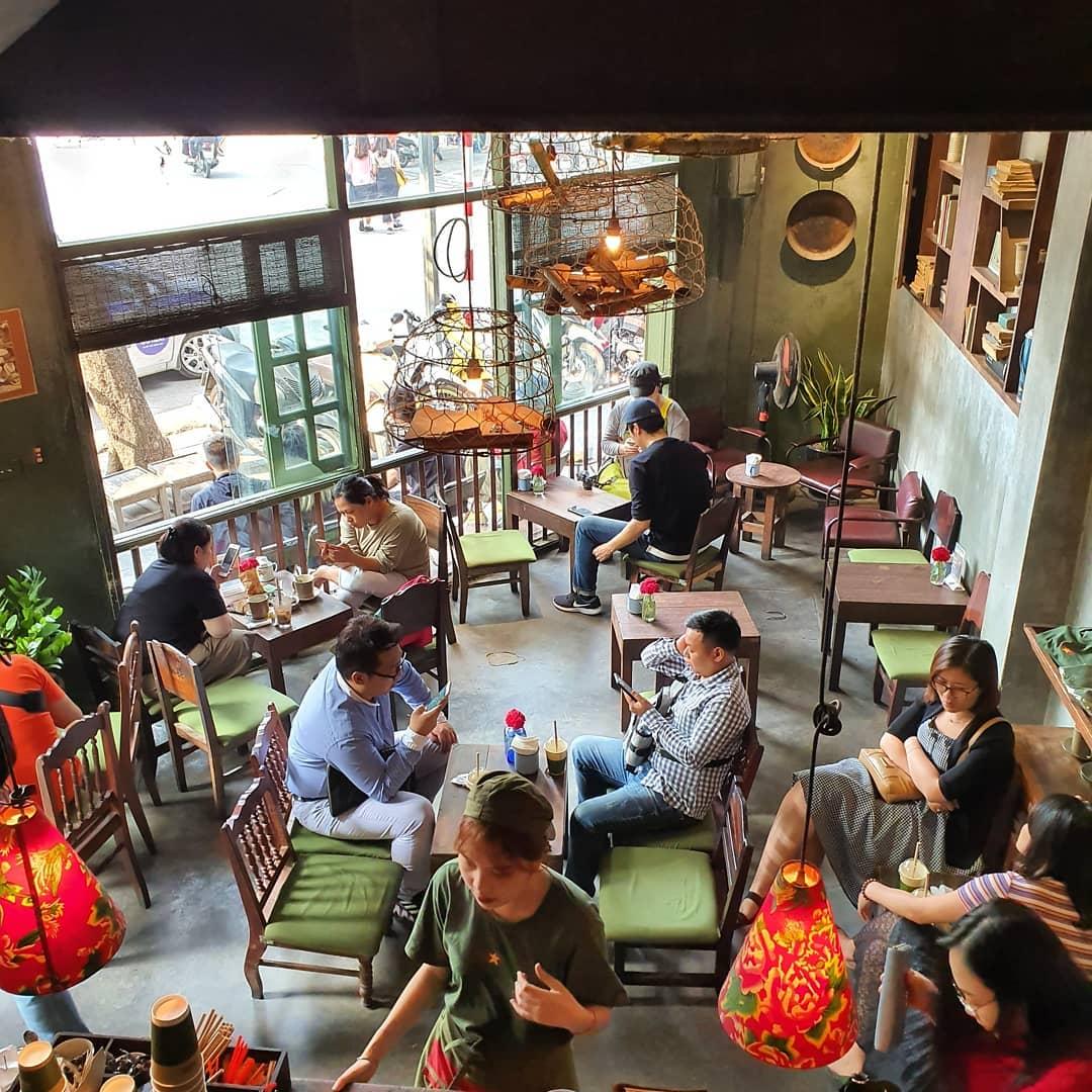 Khung cảnh phía bên trong Cộng Nhà Thờ. Vẫn là tông màu nâu – xanh rêu chủ đạo với những chiếc bàn, chiếc ghế gỗ nhỏ xinh – đặc trưng của cafe đường phố Hà Nội (Nguồn ảnh: Internet)